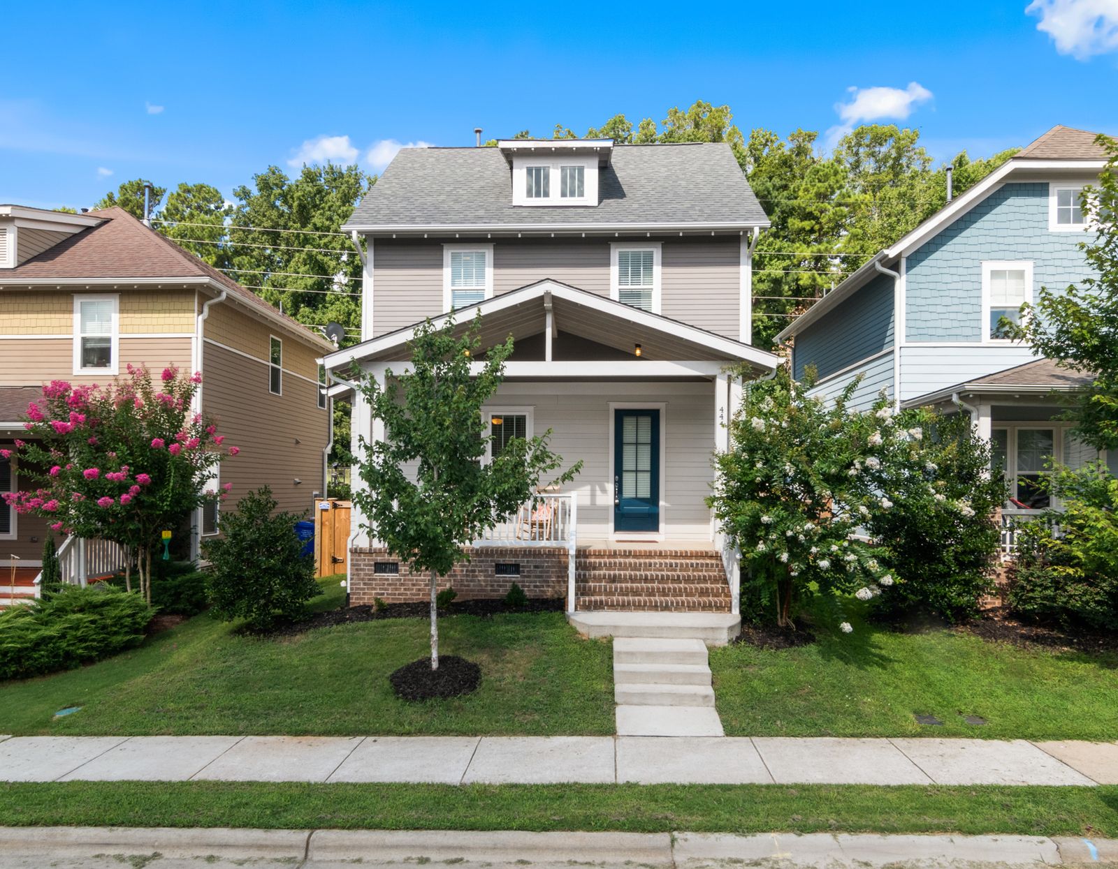 A gray porch-front home with a blue door and flowering shrubs between neighboring houses.