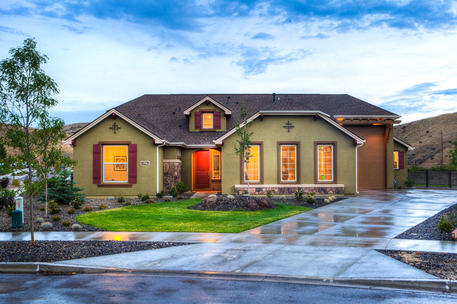 A landscaped home with warm window lights and a wet driveway at dusk.